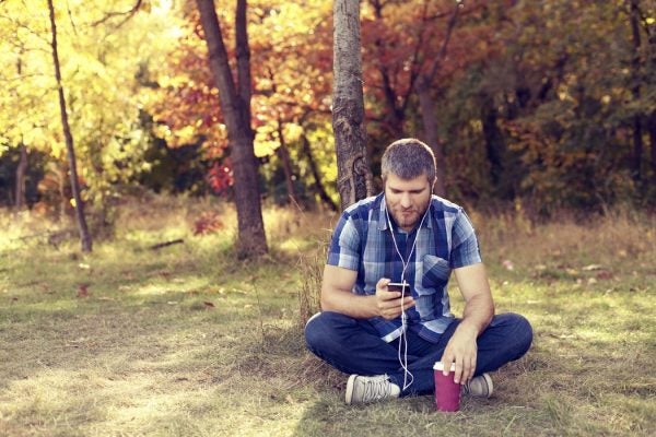 Man sitting cross-legged in a forest listening to headphones while holding a smart phone in one hand and balancing a cup of coffee in the other