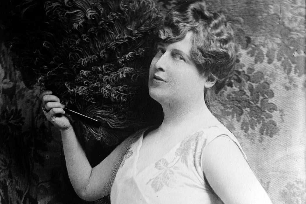 Older photograph of Florence Foster Jenkins standing agains a flowery backdrop while holding a fully feathered fan
