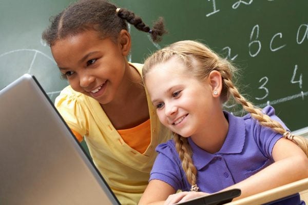 Two girls look on at a laptop together in a classroom