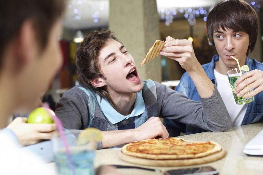 A teen looks on as his friend eats a slice of pizza