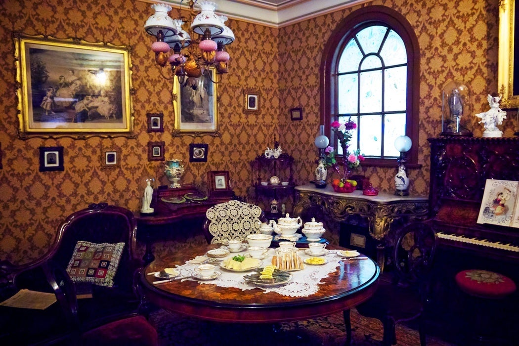 The inside of a quant Victorian parlor with mustard wallpaper, an intricately carved piano, and decor ranging from colorful flowers to vases