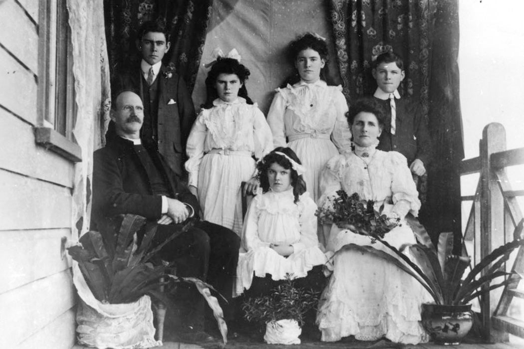 A family poses for a portrait in front of a fabric backdrop on the veranda of their home, in the early 1900s.