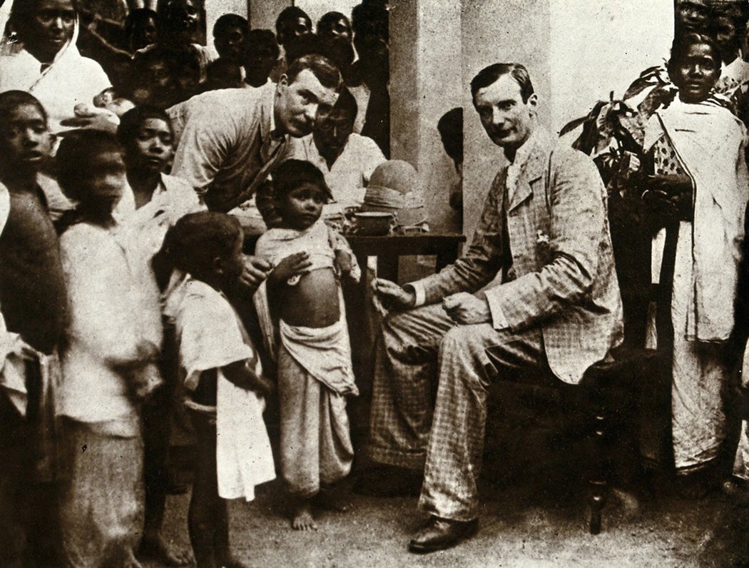 Two men in suits prepare to inoculate a child, surrounded by a crowd of parents and children. Photograph, 1880/1900.