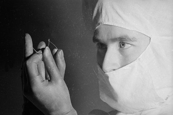 A man wearing a surgical mask and gloves threading his needle with suture before an operation.