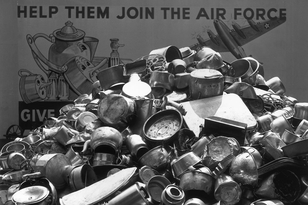 A pile of pots, pans, and kitchen utensils sits in front of a poster urging people to donate aluminum kitchen ware to help the US Air Force
