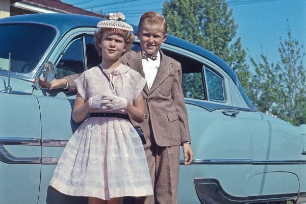 Boy and girl standing in front of camera with car.