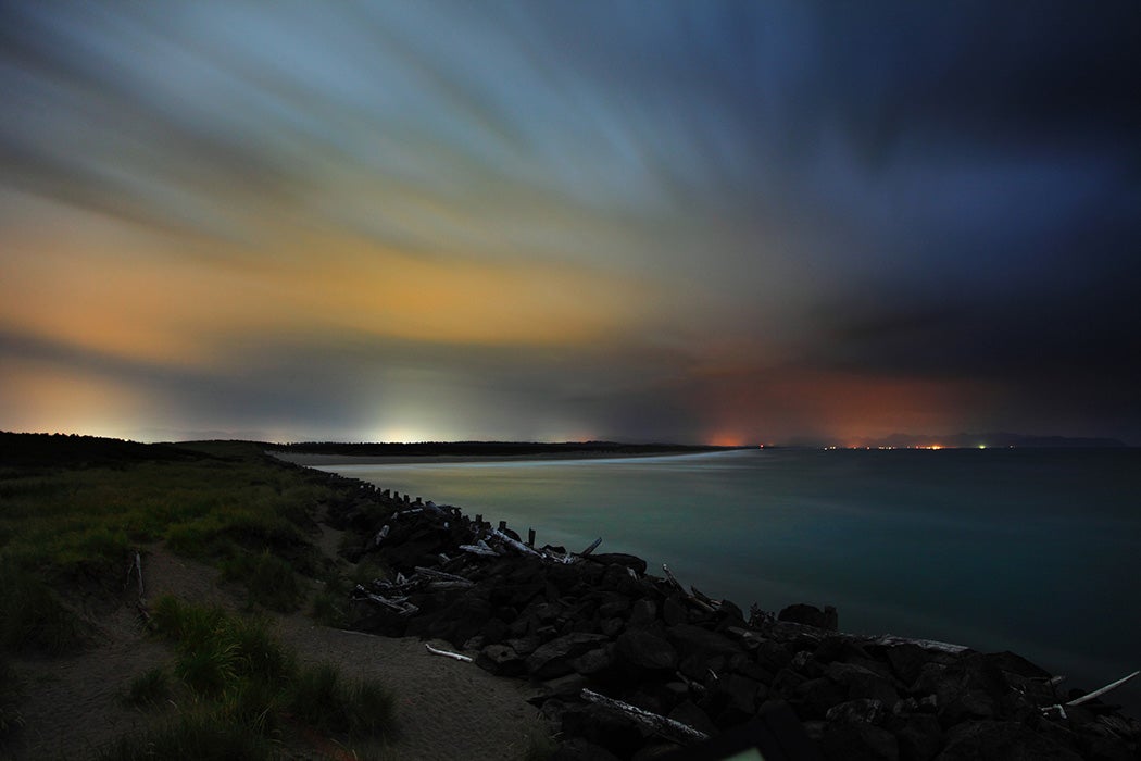 On peut voir les lumières lointaines d'Astoria, dans l'Oregon, éclairer le ciel couvert et nuageux au-dessus de la côte de l'Oregon, près de l'embouchure du fleuve Columbia