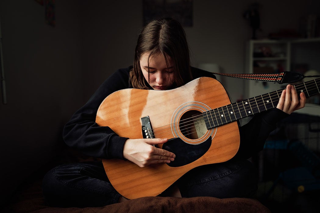 Depressed teen girl in black clothes playing guitar sitting on bed in her room.