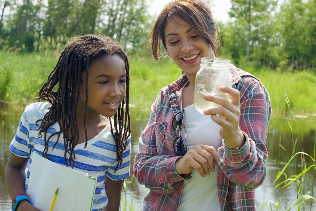 A teacher is standing next to a young student examining her findings from the pond.