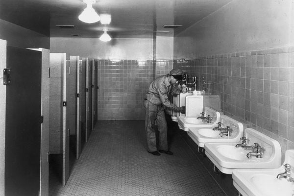 A male janitor stands and bends over a urinal in a bathroom, scrubbing the porcelain with a detergent.