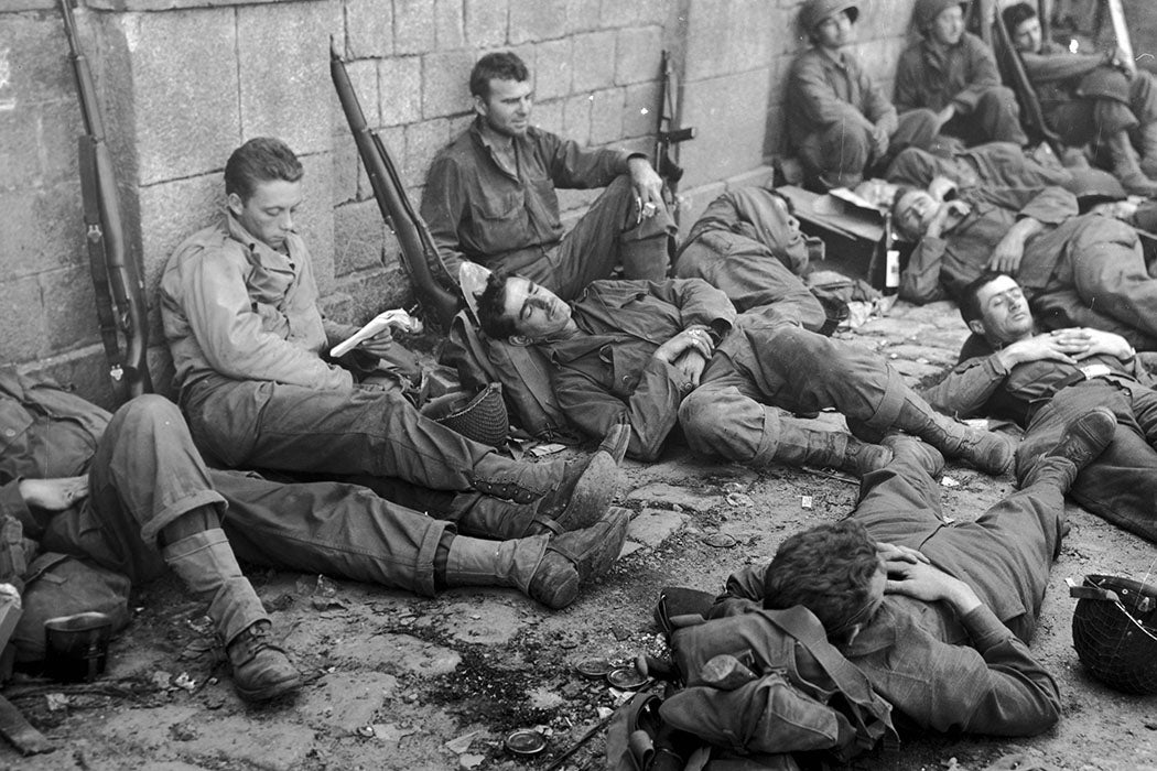 US infantrymen rest during their drive to follow armored units south from Normandy into Brittany, Villedieu, France. Some men sit and lean their rifles against a stone wall, while others lay on the ground, resting their heads on their backpacks.
