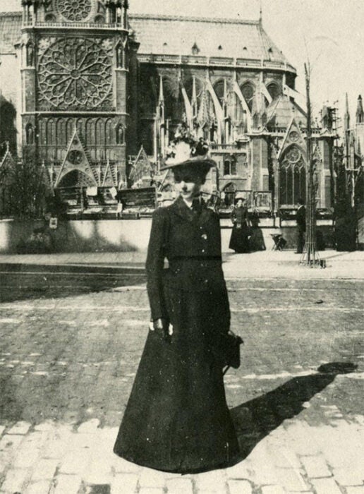 Architect Julia Morgan in Paris standing in front of the Notre Dame. Black and white image of Morgan in long black dress with elaborate hat.