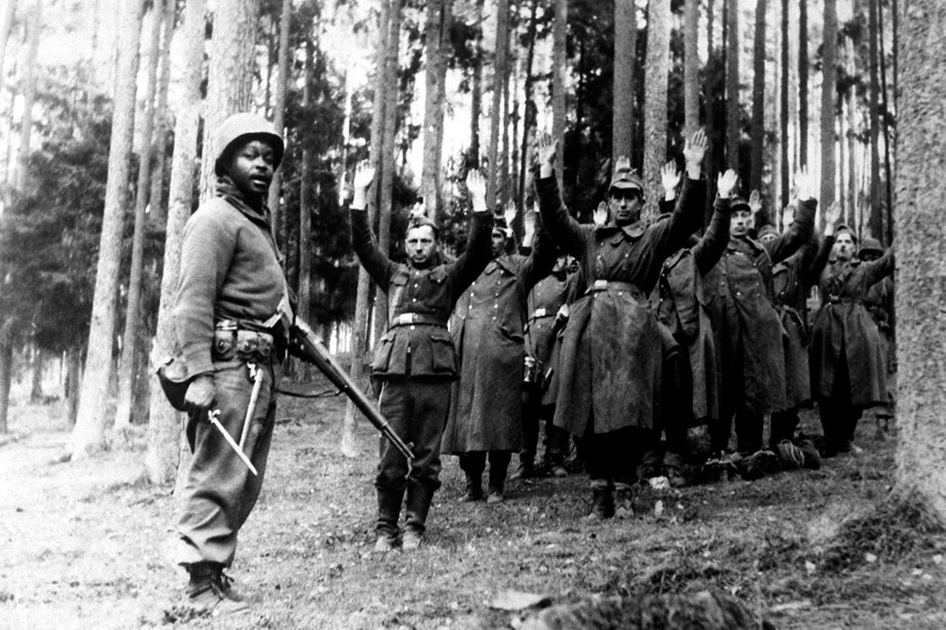 A Black soldier of the 12th Armored Division stands guard over a group of Nazi prisoners captured in the surrounding German forest, April 1945