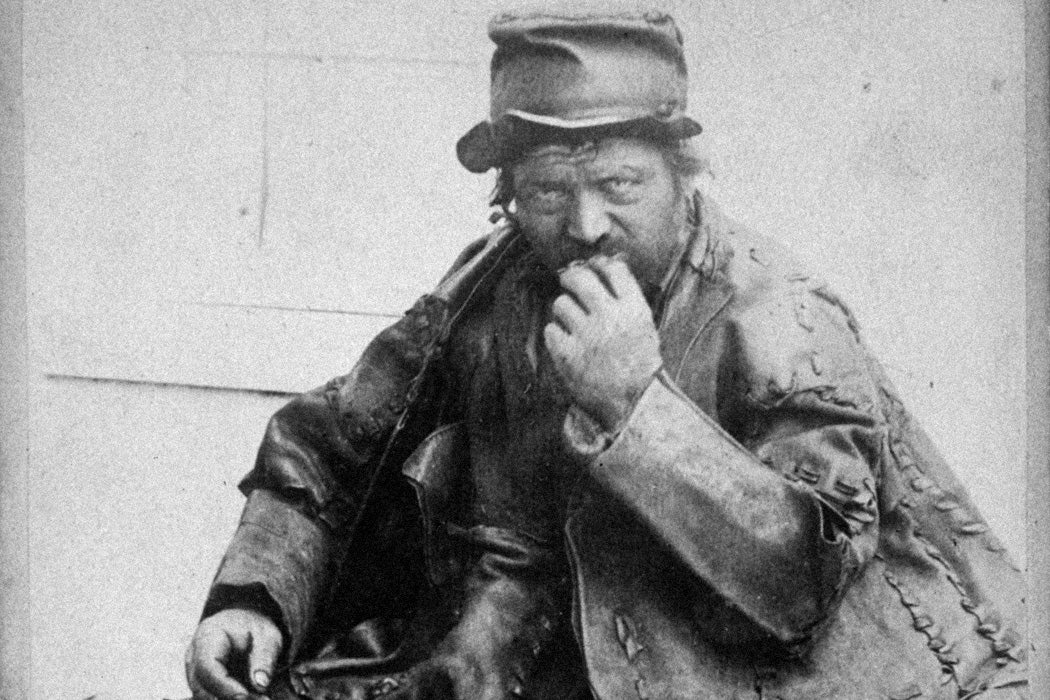 An aged photo of the Leatherman wearing a thick leather coat and leather pants, looking at the camera while sitting and eating, circa June 9, 1885