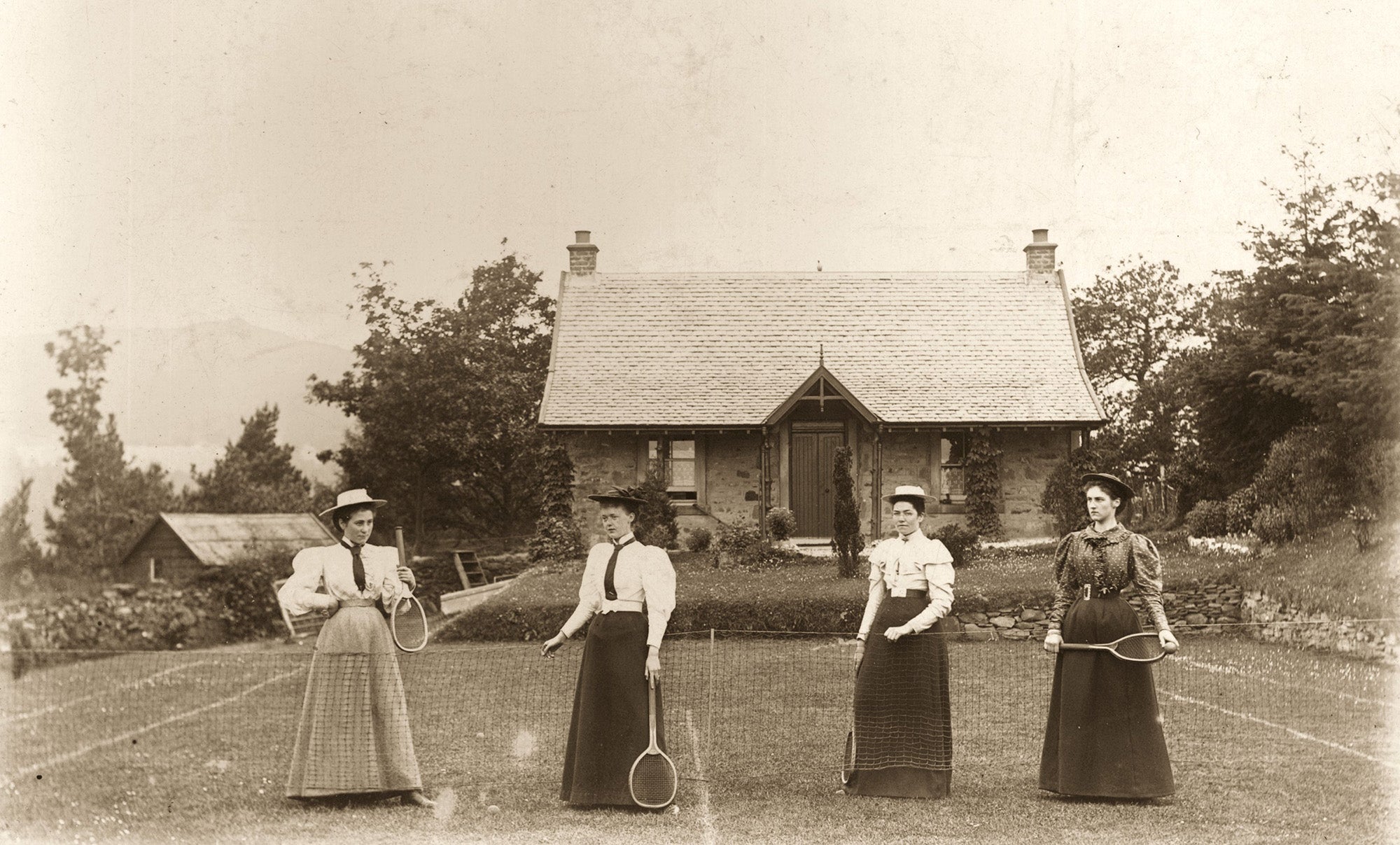 Four Victorian ladies in full-length skirts prepare for a game of lawn tennis.