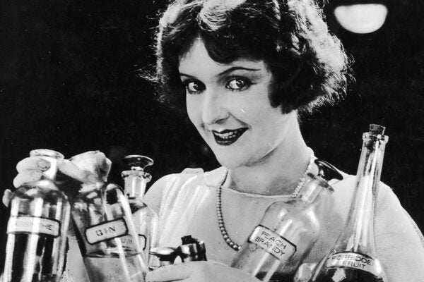 A woman smiles while holding bottles of various types of alcohol, including peach brandy, port wine, gin, absinthe, and forbidden fruit.
