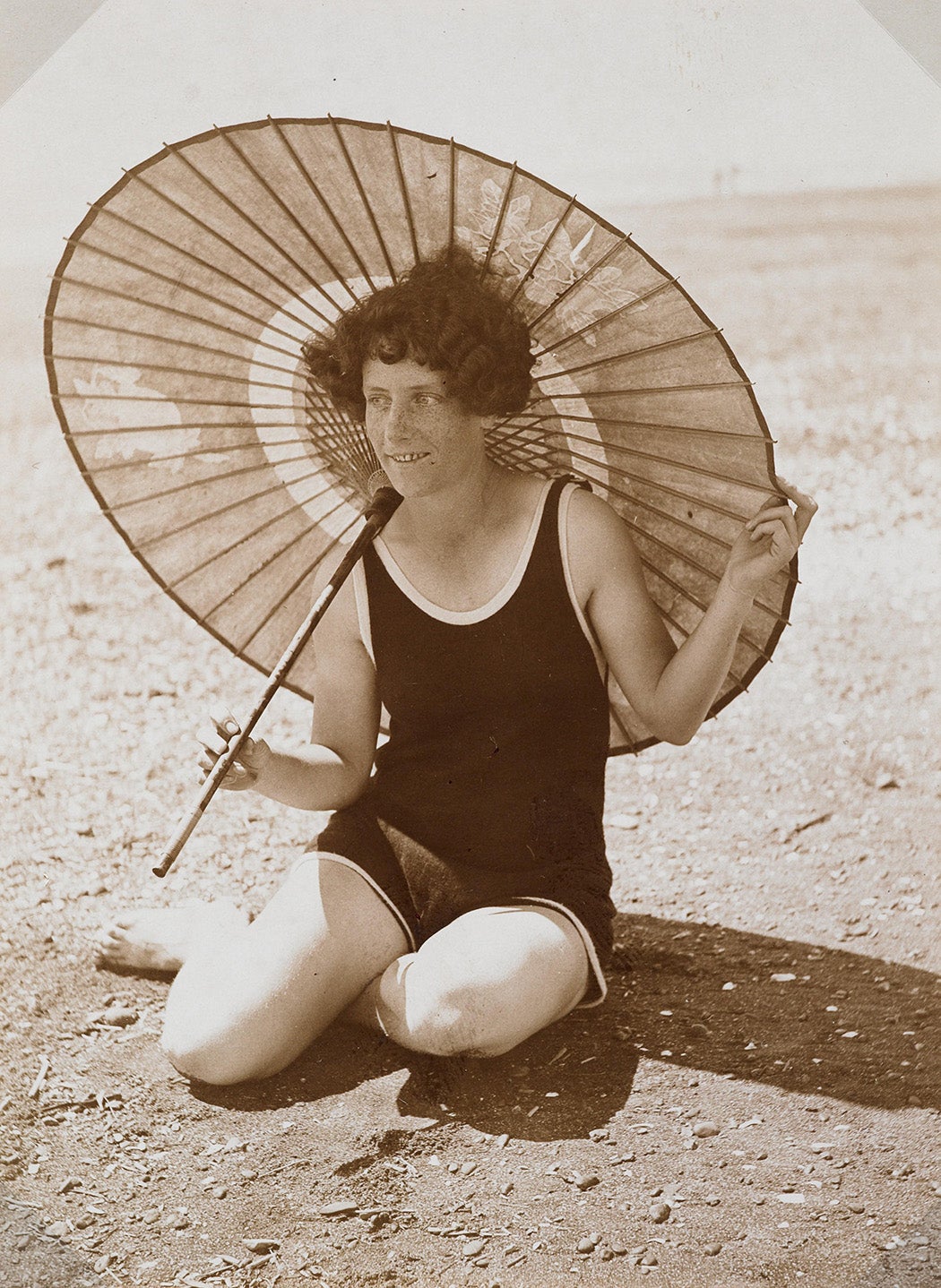 A woman in a dark bathing suit sitting on a beach, holding a Chinese-style parasol over her shoulder. 