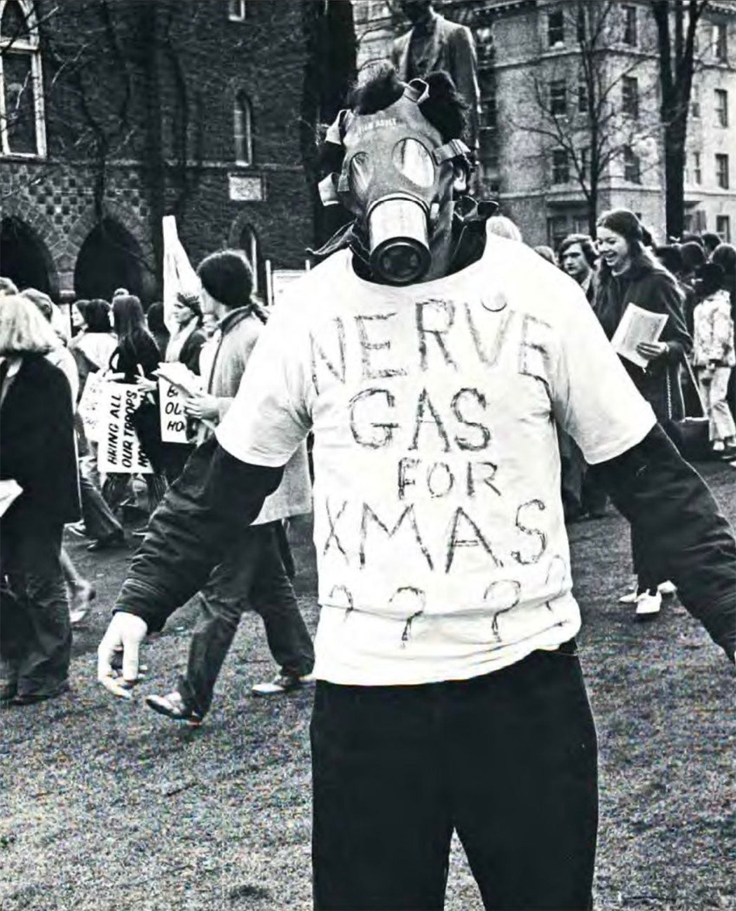 A black and white image of a man facing the camera, wearing a gas mask and a shirt that reads "Nerve gas for xmas????"