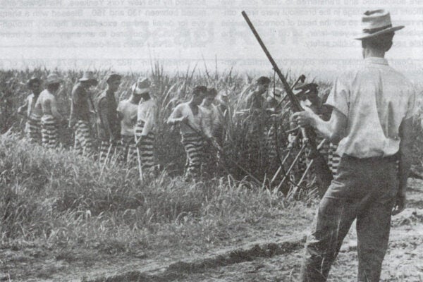 A page from The Angolite that features a photograph of a prison guard holding a shotgun while watching prisoners work in a field.