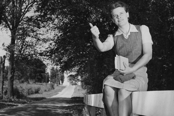 A woman sitting on a fence, books in her left hand while thumbing a lift with her right hand, on a country road, United States, circa 1955.