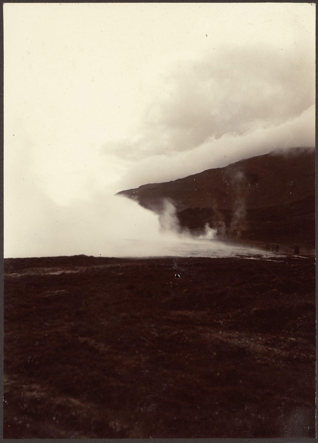 People watching an erupting geyser. Water and stream rising in the air, mountains in background.