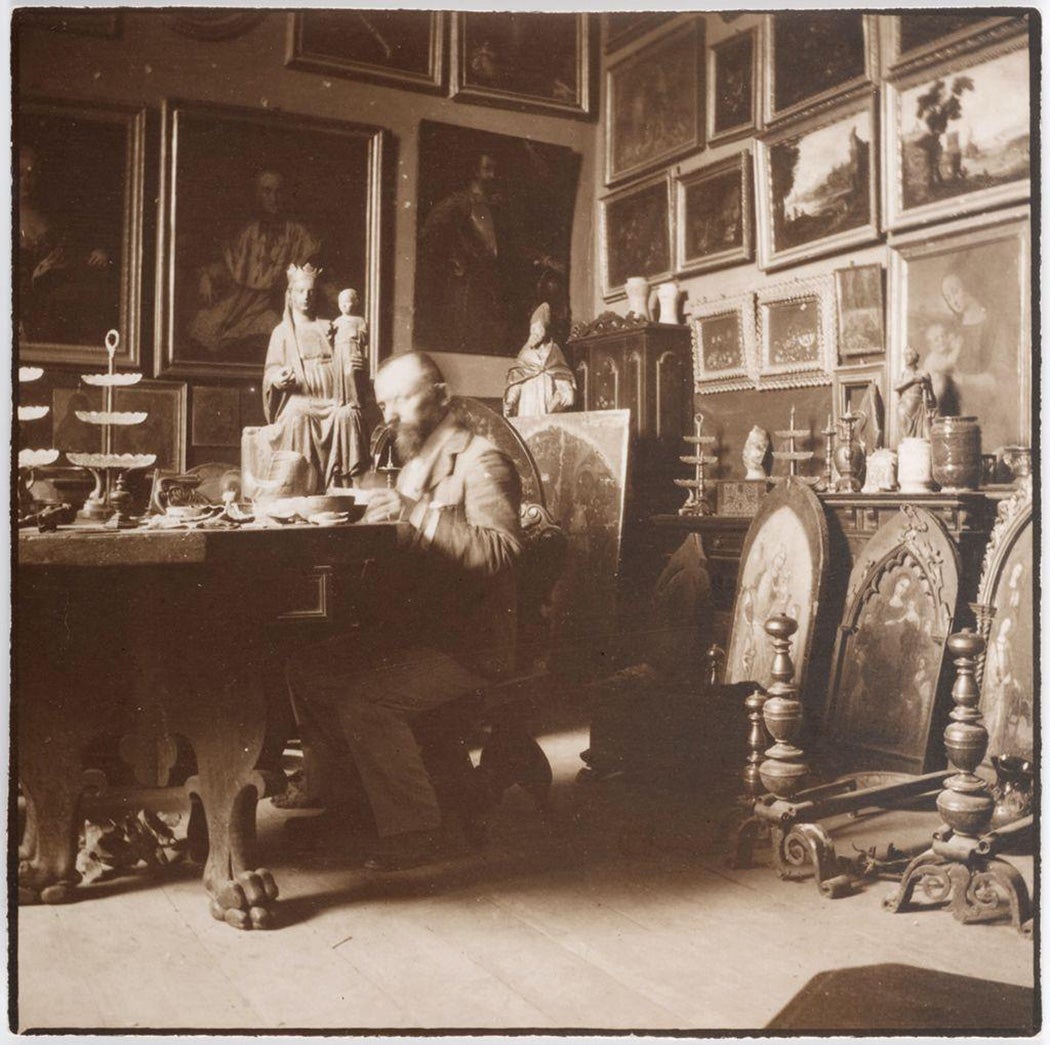 Bernard Berenson sitting at a table in a room filled with paintings. 