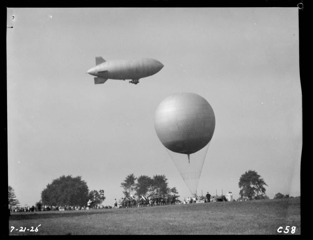 A band plays next to a balloon while Goodyear's blimp, Pilgrim, soars overhead, July 1925