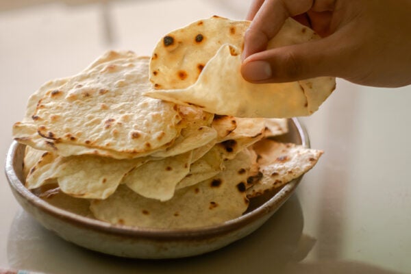 a close-up of a hand selecting a freshly made roti from a stack on a rustic ceramic plate. The warm and natural lighting emphasizes the texture and golden-brown spots on the roti, making it an appealing visual for themes related to traditional cuisine.