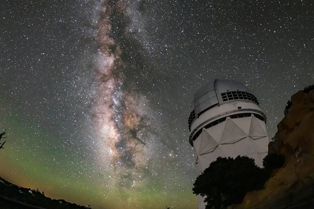 A white structure with a domed top on a rocky outcrop below a star-filled night sky. The Milky Way stretches vertically in a luminous band. Faint green glow tints the curved horizon.