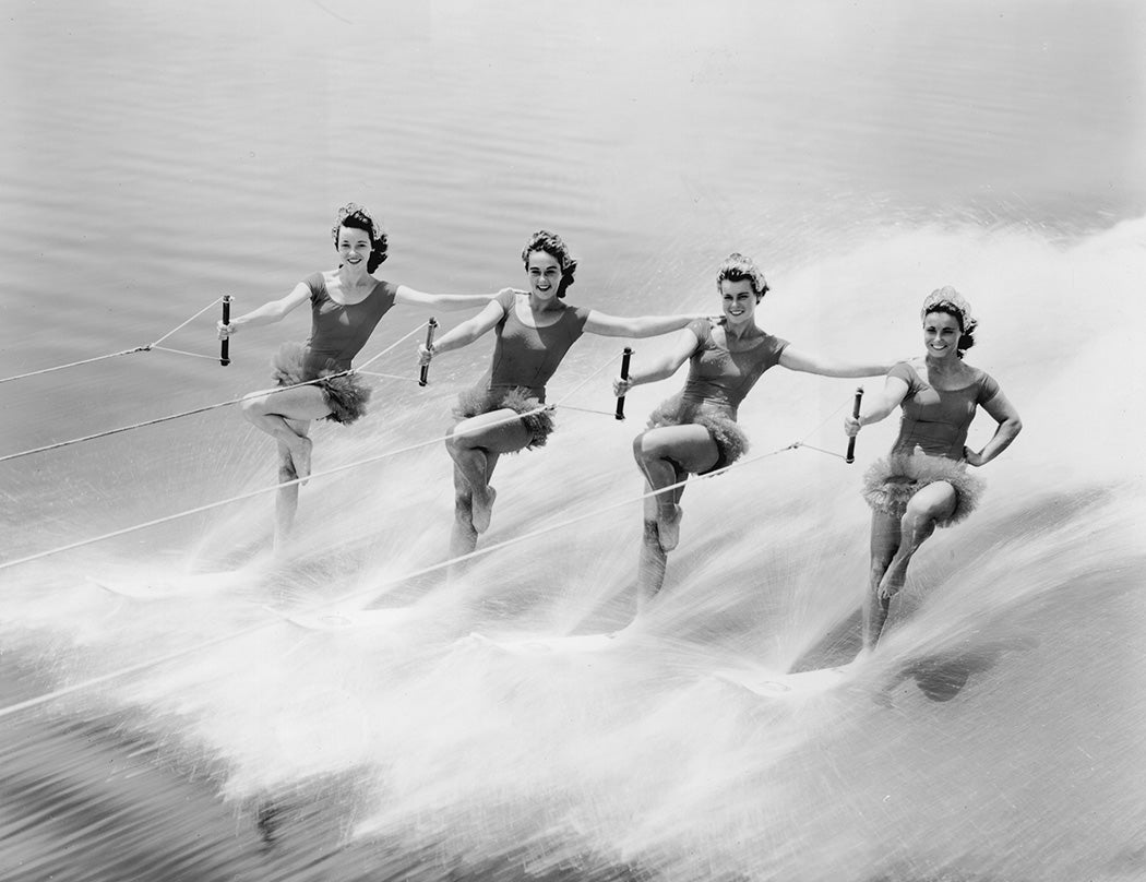 Four female water skiers perform tricks; each rests one arm on another's shoulder and stands on one ski.