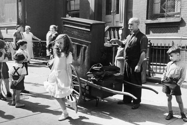 An organ grinder stands on a sidewalk, playing music as a young girl dances in front of him, New York City, ca. 1935