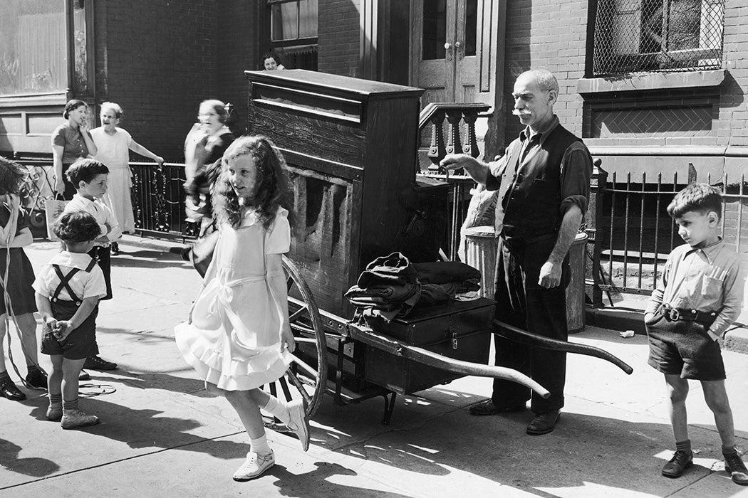An organ grinder stands on a sidewalk, playing music as a young girl dances in front of him, New York City, ca. 1935