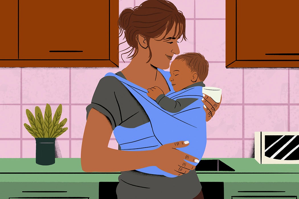 Smiling woman holding a sleeping baby in a blue sling in a cozy kitchen setting