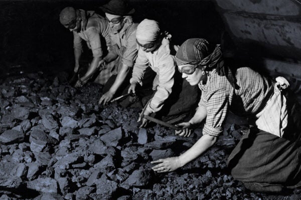 Women crack coal wearing goggles, using their bare hands and tiny hammers in the damp darkness of a tunnel in a mine.
